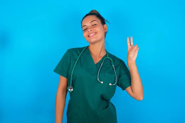 青い背景の上に緑色の制服を着た美しいヒスパニック系の医者の女性が勝利のサインをしてカメラに顔をウィンクして笑っています 第二番 — ストック写真