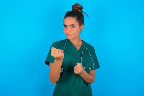 青い背景に緑の制服を着た美しいヒスパニック系の医師の女性拳の防衛ジェスチャーで戦う準備ができて 怒りと動揺顔 問題を恐れて — ストック写真