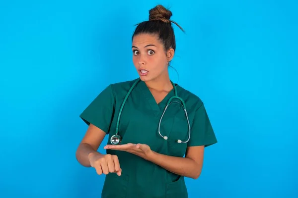 青い背景の上に緑の制服を着た美しいヒスパニック系の医師の女性が急いで時間を見て指して 動揺し 締め切り遅延のために怒っています — ストック写真