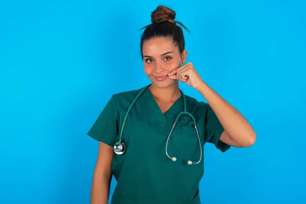 青い背景の口と唇に緑色の制服を着た美しいヒスパニック系の医師の女性が指でZipとしてシャットダウンします 秘密裏に黙ってタブーを語る — ストック写真