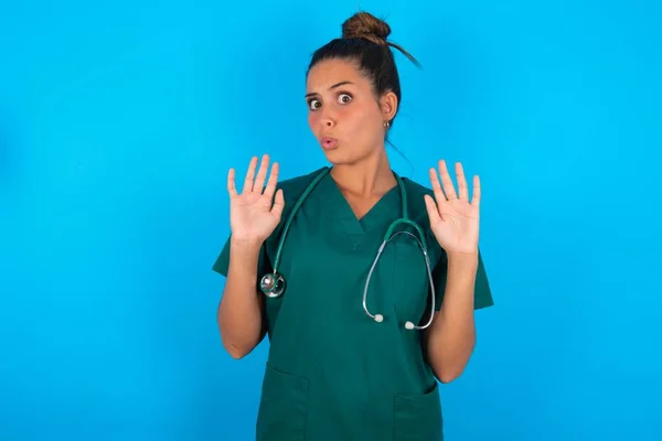 青い背景に緑の制服を着た美しいヒスパニック系の医師の女性恐怖と嫌な表情で拒否と否定を示す手のひらを離れて移動します 止めて禁止 — ストック写真