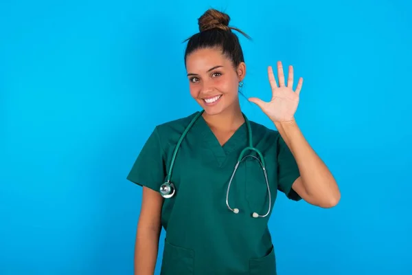 青い背景の上に緑色の制服を着た美しいヒスパニック系の医者の女性が自信と幸せ笑顔ながら指の数5で見上げと指摘 — ストック写真
