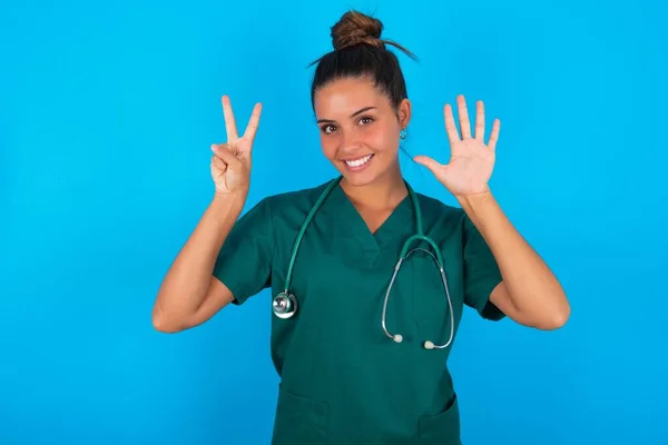 青い背景に緑色の制服を着た美しいヒスパニック系の医者の女性が自信を持って幸せな笑顔で指の数7を示して指摘しながら — ストック写真