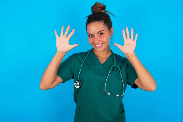 自信満々で笑顔で指10番を見せ指差す青い背景に緑の制服を着た美しいヒスパニック系の医者の女性 — ストック写真