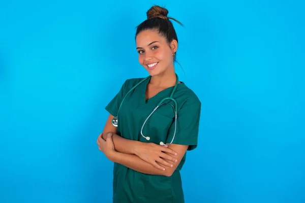 青い背景の上に緑色の制服を着た美しいヒスパニック系の医者の女性カメラを見て交差腕で笑顔幸せな顔 正の人 — ストック写真