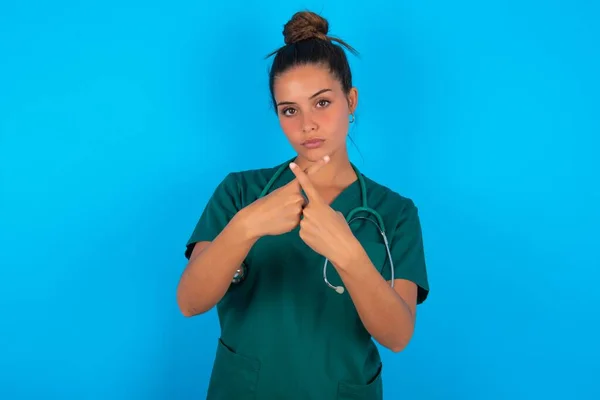 青い背景に緑の制服を着た美しいヒスパニック系の医師の女性が否定的な記号を行う指を横断拒絶怒りの式を持っています — ストック写真