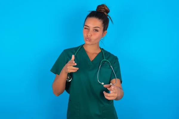 喜びの美しいヒスパニック系の医師の女性を身に着けています緑の医療制服上の青の背景ウィンクとポイントインデックス指でカメラ 誰かを選択します — ストック写真