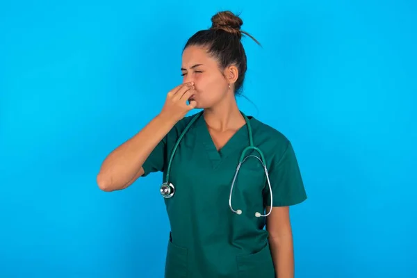 青い背景に緑色の制服を着た美しいヒスパニック系の医者の女性は臭いと嫌な匂いを嗅ぎ鼻に指で息をしています 匂いが悪い — ストック写真