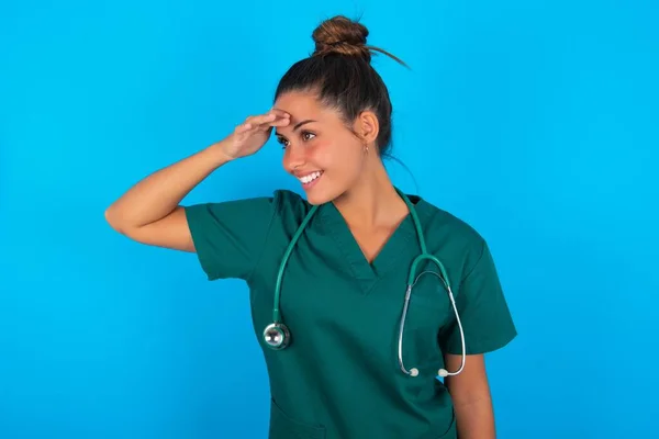 青い背景に緑色の制服を着た美しいヒスパニック系の医師の女性は非常に幸せと笑顔遠くに手を頭の上に見て 検索の概念 — ストック写真