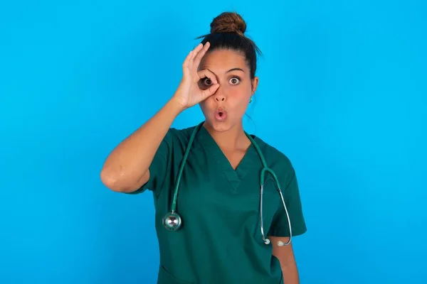 青い背景に緑色の制服を着た美しいヒスパニック系の医師の女性は 驚きの顔でショックを受けたOkジェスチャーを行うと 目の指を見て 不信仰の表現 — ストック写真