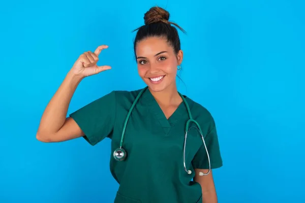 青い背景に緑色の制服を着た美しいヒスパニック系の医者の女性が笑顔で 指を見てカメラで小さなサイズの看板をして手で自信を持ってジェスチャー 概念の測定 — ストック写真