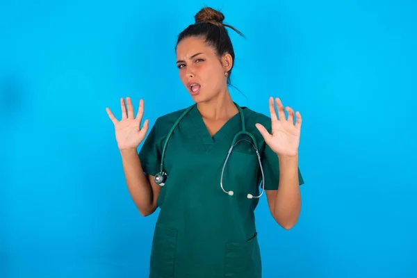 青い背景の上に緑色の制服を着た美しいヒスパニック系の医者の女性は恐怖の表情で恐れて手でジェスチャーを停止し ショックで叫んでいます パニック概念 — ストック写真