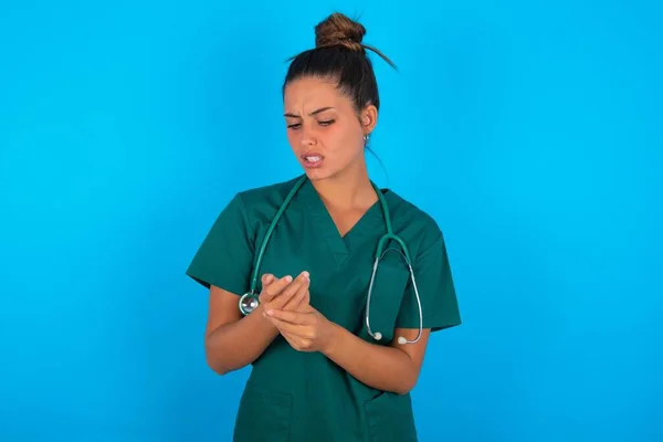 青い背景に緑の制服を着た美しいヒスパニック系の医師の女性手や指の痛み 関節炎の炎症 — ストック写真