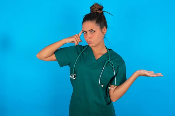 青い背景の上に緑色の制服を着た美しいヒスパニック系の医師の女性が混乱し コピースペースを示し 額に指を指すオープンヤシに悩まされています 考えてみて — ストック写真