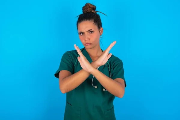 青い背景に緑の制服を着た美しいヒスパニック系の医師の女性否定的な記号を行う腕を横断拒否式 怒りの顔 — ストック写真