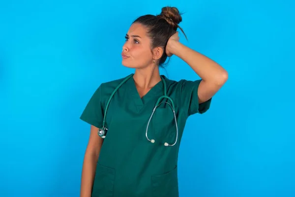 青い背景の上に緑色の制服を着た美しいヒスパニック系の医師の女性が混乱し 疑問に思います 疑いもなく 頭の上で手で考えて 包括的概念 — ストック写真