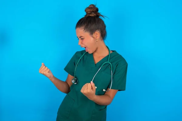 青い背景の上に緑の制服を着た美しいヒスパニック系の医師の女性は非常に幸せと腕を上げて勝者のジェスチャーを行う興奮 笑顔と成功のために叫んで お祝いのコンセプト — ストック写真