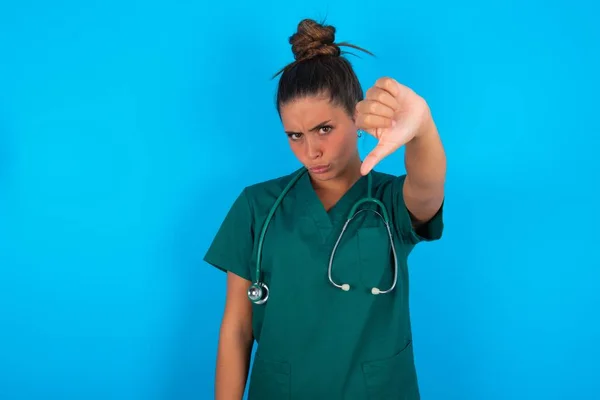 青い背景色の上に緑色の制服を着た美しいヒスパニック系の医師の女性が怒り いらいら 失望または不満を感じ 深刻な外観で親指を下に示す — ストック写真