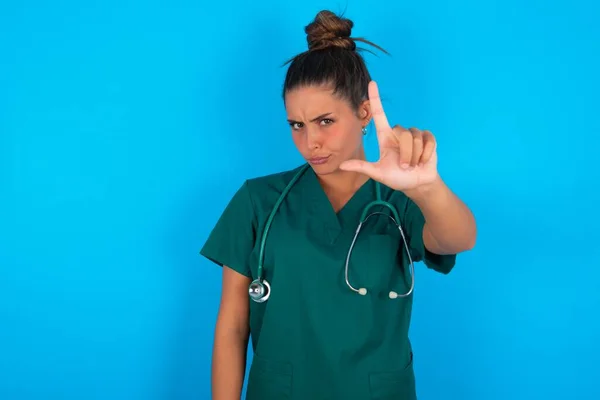 美しいですヒスパニック医師女性身に着けています緑医療制服上の青の背景作り楽しいです人々とともに指で額を行います負け犬のジェスチャーモックと侮辱 — ストック写真