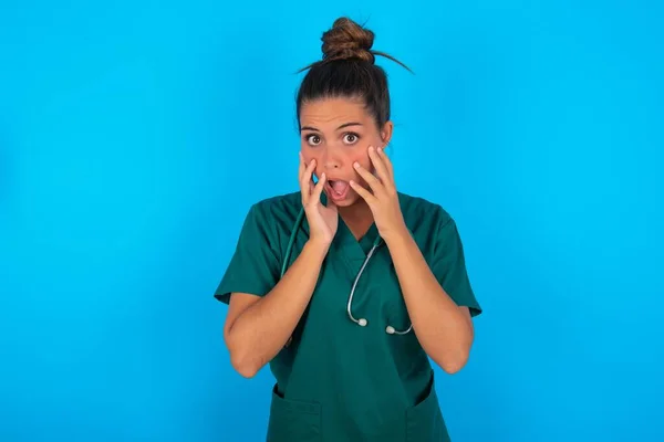 青い背景に緑の制服を着た美しいヒスパニック系の医師の女性が興奮とスリルを表現し 顎を落とし続け 頬に手を出し 目が出てきました — ストック写真