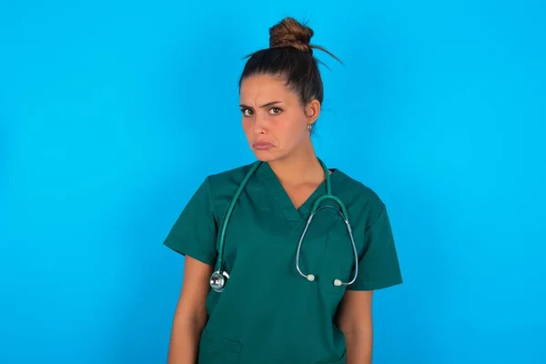 青い背景の眉の上に緑の医療制服を身に着けている不幸な動揺美しいパニック医師の女性は泣きます 目標を達成することができないとして不満と不幸されています 失望モデルは悩みを持っています — ストック写真