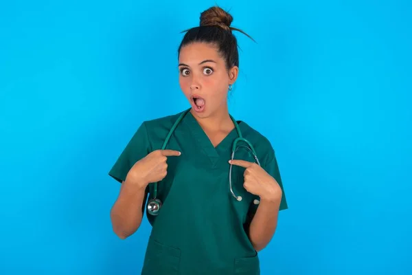Peinlich Schöne Hispanische Ärztin Grüner Arztuniform Vor Blauem Hintergrund Zeigt — Stockfoto