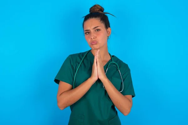 青い背景の笑顔の上に緑の医療制服を着た積極的な美しいパニック医師の女性は喜んで 対話者から楽しいニュースを受け取るために喜んで 一緒に手のひらを保持し 人々の感情の概念 — ストック写真