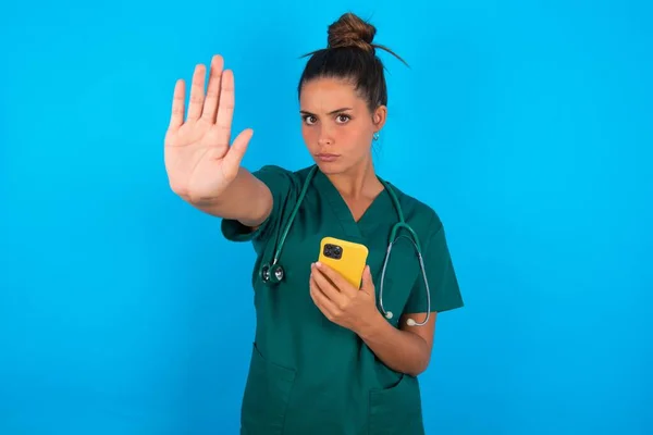青の背景に緑の制服を着た美しいヒスパニック系の医師の女性が使用して 真剣に自信を持って表現 防衛ジェスチャーで停止標識を行うオープン手でスマートフォンでテキストメッセージ — ストック写真