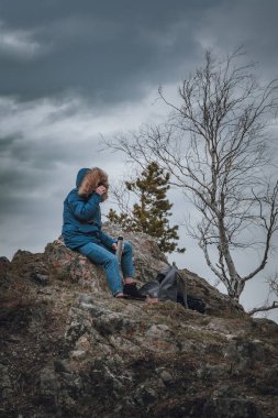 Young woman in a blue down jacket drinks tea from a thermos on a mountain top under a gloomy gray sky. High quality photo
