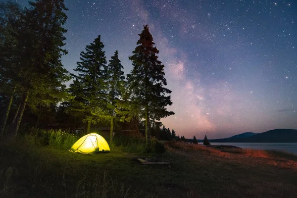 Milky Way and sky full of stars above the luminous tent under the trees by the lake — Foto Stock