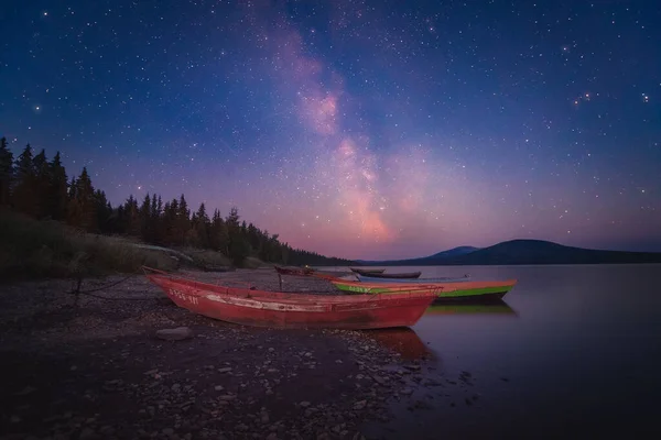 Milky Way and sky full of stars above the lake and moored boats with mountains on the horizon — стоковое фото