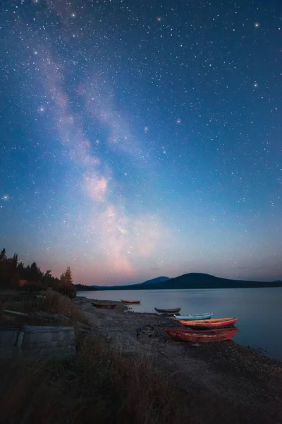 Milky Way and sky full of stars above the lake and moored boats with mountains on the horizon — Stok fotoğraf