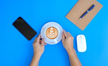 woman hand holding cup of coffee and smartphone on blue background, top view