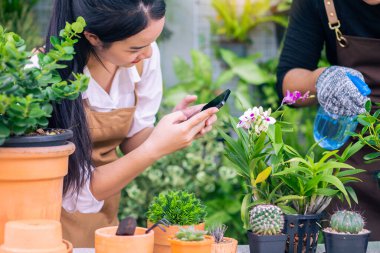 Happily Asian young gardener couple wearing apron use garden equipment and laptop computer to take care the house plants in greenhouse shop together, small business with green plant