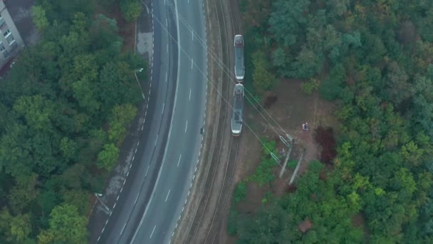 路面電車は霧の多い夏に緑の森の近くのレールで街を旅します 上からの眺めを追跡します 市内の鉄道で2台の車に乗るトラム ドローンのオーバーヘッドショットの追跡 — ストック動画