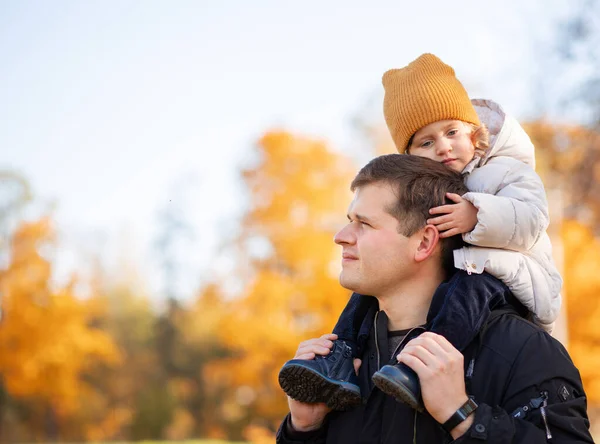 Father gives his son a ride on his back in the park. Portrait of a pensive father giving his daughter a piggyback ride on his shoulders and looking up. Cute baby girl with her father