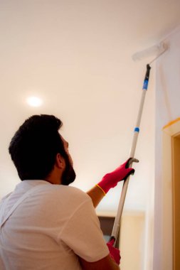 Koyu saçlı, sakallı, turuncu bir duvarı yuvarlak silindirli beyaz boyayla boyayan ve duvarın üst kısımlarına daha iyi erişmek ve koruyucu eldiven takmak için genişletici kullanan bir adam..