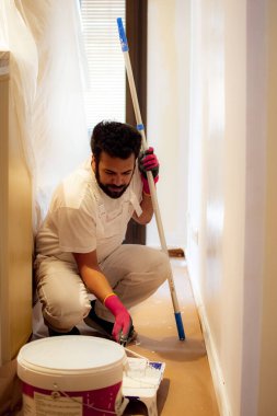 Koyu saçlı, sakallı, turuncu bir duvarı yuvarlak silindirli beyaz boyayla boyayan ve duvarın üst kısımlarına daha iyi erişmek ve koruyucu eldiven takmak için genişletici kullanan bir adam..