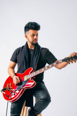 Siyah saçlı, sakallı, kahverengi tenli, siyah elbiseli, tahta bir taburede oturan ve beyaz arka planda kırmızı elektro gitar tutan Latin bir adamın stüdyo fotoğrafı..