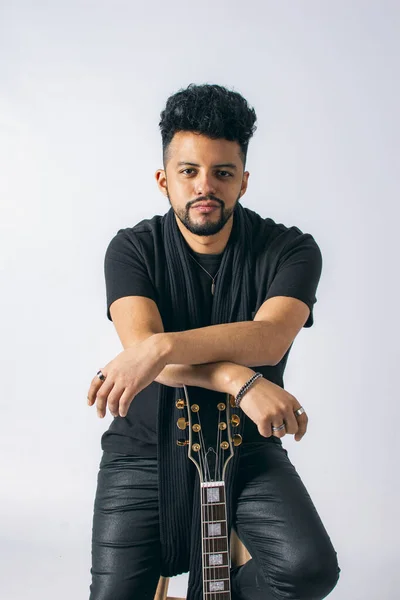 Koyu tenli ve sakallı Latin bir adamın portresi, siyah giyinmiş, beyaz arka planda kırmızı bir elektro gitara yaslanmış..