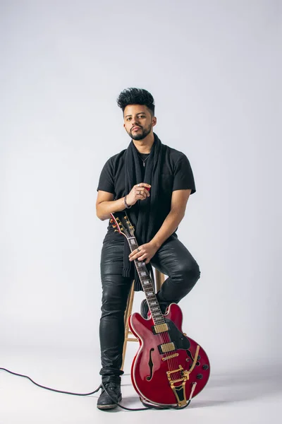Siyah saçlı, sakallı, kahverengi tenli, siyah elbiseli, tahta bir taburede oturan ve beyaz arka planda kırmızı elektro gitar tutan Latin bir adamın stüdyo fotoğrafı..
