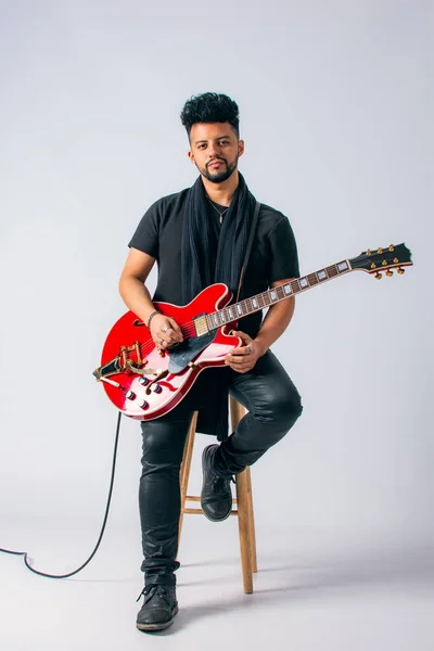 Siyah saçlı, sakallı, kahverengi tenli, siyah elbiseli, tahta bir taburede oturan ve beyaz arka planda kırmızı elektro gitar tutan Latin bir adamın stüdyo fotoğrafı..