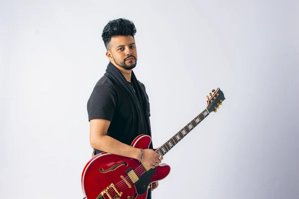 Esmer, siyah saçlı ve sakallı, beyaz arka planda kırmızı elektro gitar çalan Latin bir adamın stüdyo fotoğrafı..