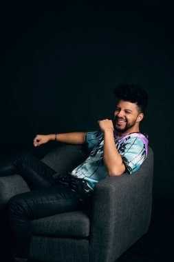 Koyu renk saçlı ve sakallı, siyah arka planda renkli bir gömlek giymiş, gri koltukta oturan Latin bir adam..