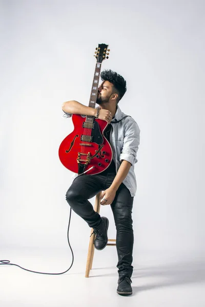 Koyu renk saçlı, sakallı, tahta taburede oturan, kot ceket giyen ve beyaz arka planda kırmızı elektro gitar tutan kahverengi tenli Latin bir adamın stüdyo fotoğrafı..