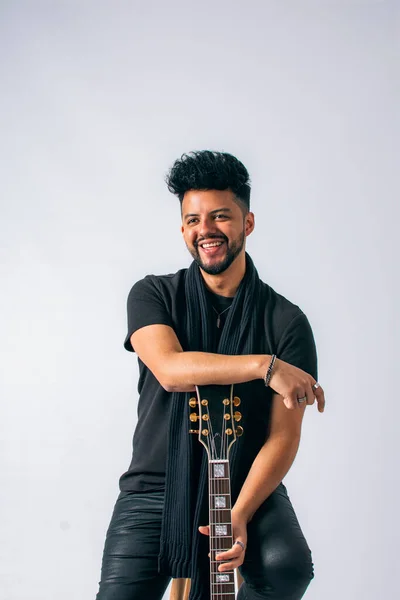 Koyu tenli ve sakallı bir Latin 'in portresi, siyah giyinmiş, kırmızı elektro gitara yaslanmış, beyaz arka planda gülüyor..