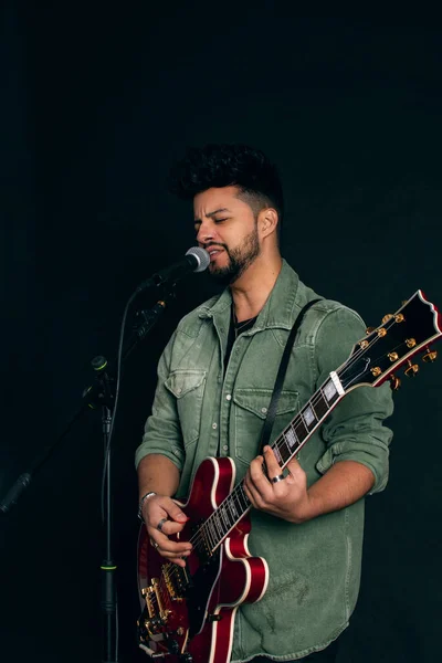 Latin bir adam şarkı söylüyor, siyah saçlı, sakallı, kırmızı elektro gitarlı ve mikrofonlu, siyah arka planda yeşil ceketli..