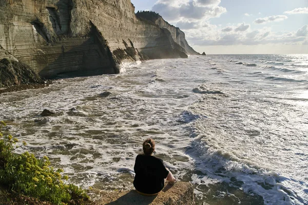 Νεαρή γυναίκα κάθεται σε μια πέτρα και θαυμάζει τη θάλασσα και τους βράχους — Φωτογραφία Αρχείου