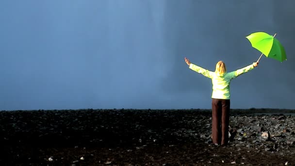 Konzeptaufnahme einer einsamen Frau, die am Rande eines Wasserfalls mit grünem Regenschirm steht — Stockvideo