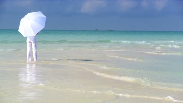 Serene jonge vrouw met een witte parasol in het ondiepe water naast een prachtige witte zandstrand — Stockvideo